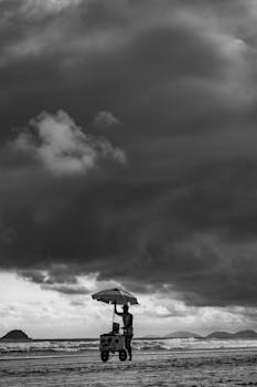 Grayscale Photo of a Man Pushing a Cart while Walking on the Shore of the Beach